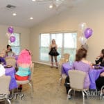 Women dressed in costumes sitting around tables with purple tablecloth. There are pearl and purple balloons and a woman stands in the middle of the room speaking