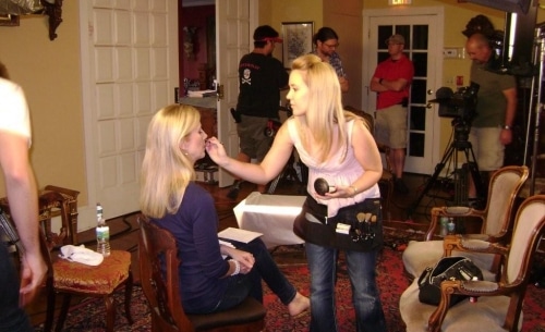 A blonde woman is applying makeup to the face of a seated blonde woman