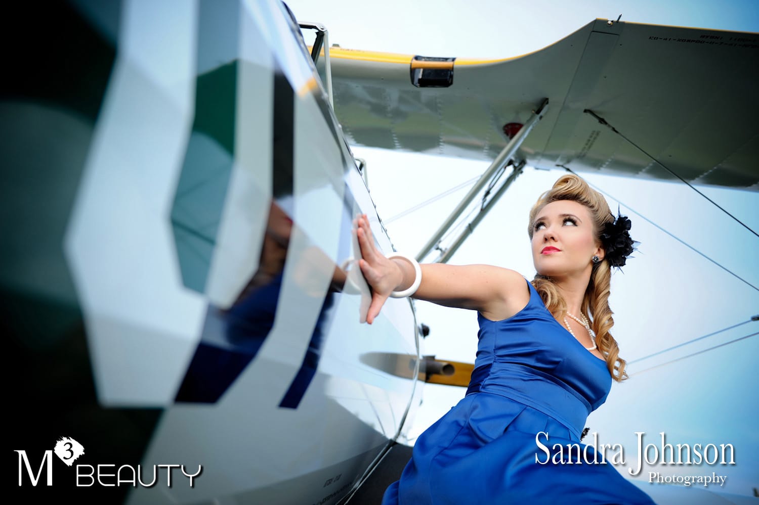Woman leaning against an airplane. She is dressed in a vintage blue dress, has blonde hair, and a black flower in her hair