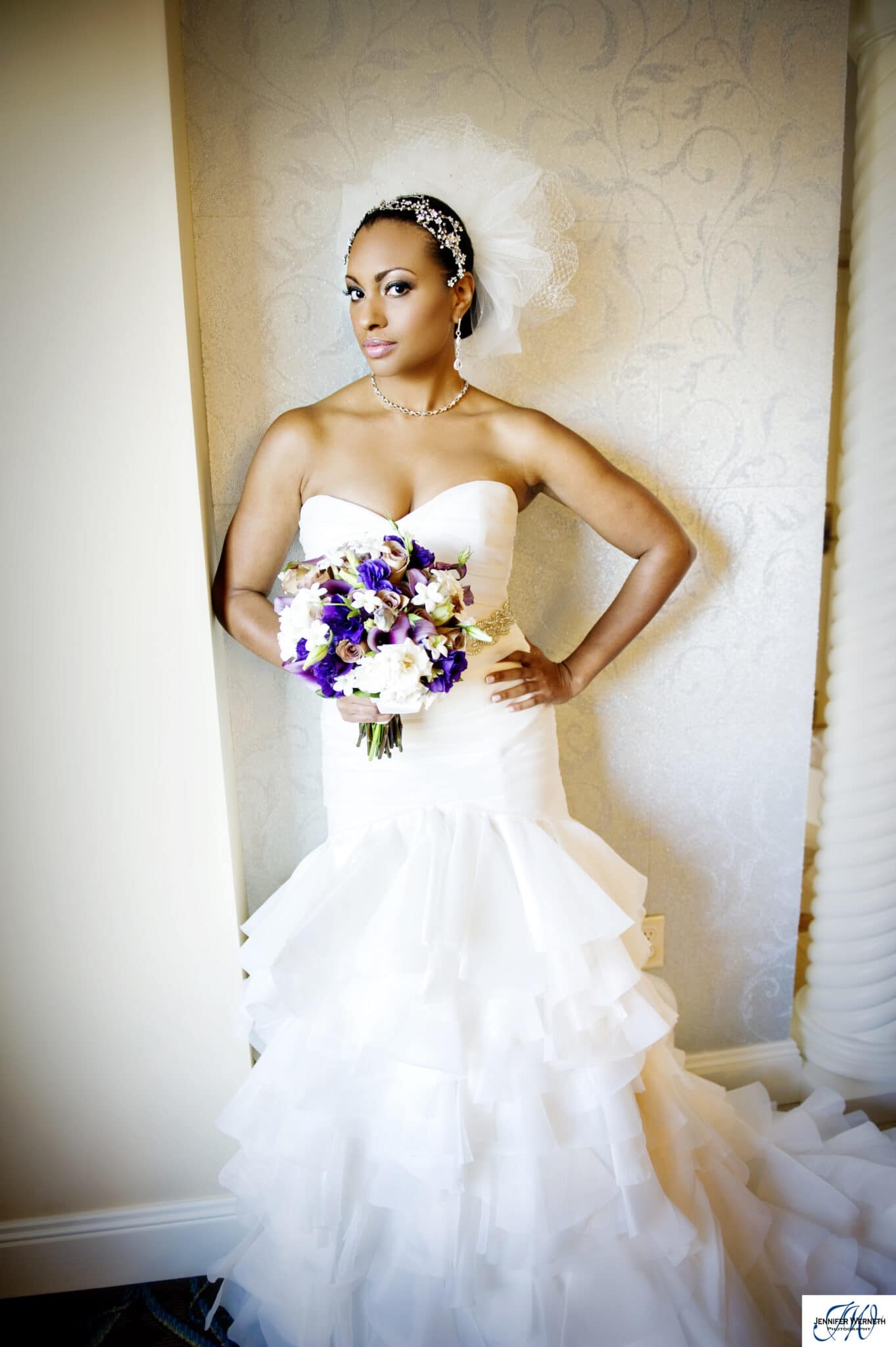 Bride leaning against a wall. She has a sleeveless dress and holds purple and white flowers