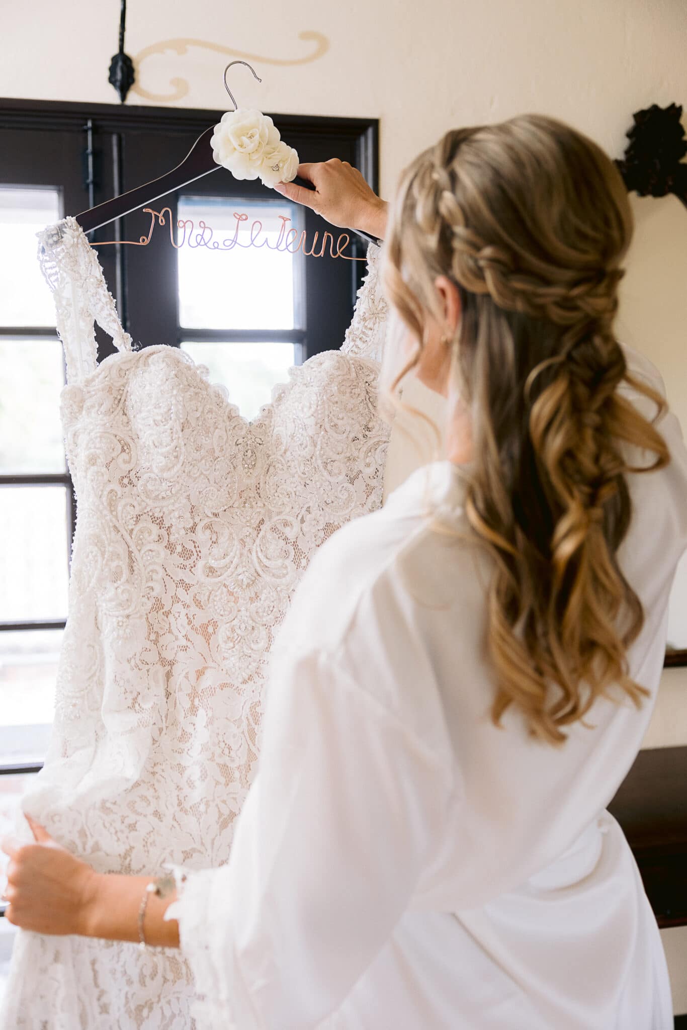 Bride facing away from the camera. Her hair is braided and down and she is taking her wedding dress off of the hanger
